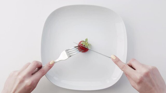 Female hands with a fork and knife cutting one strawberry on a large white empty plate. Minimalism concept Isolated on white background. Slow motion