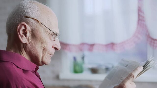 Gray haired senior man wearing glasses reading newspaper sitting at home side view