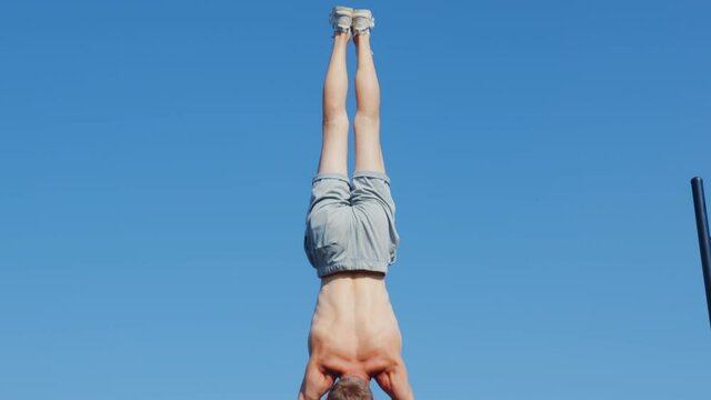 A man does a street workout at a sports ground. A sportsman does a handstand on parallel bars against an urban landscape
