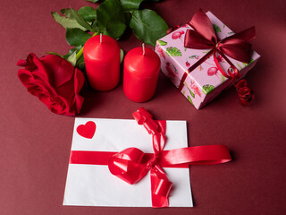A large red rose that is next to two red large candles and a pink gift