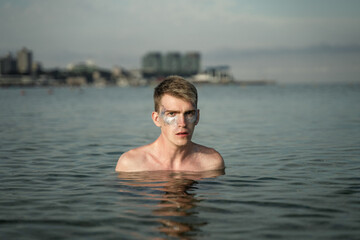 portrait of a non-binary young man with sparkles on his face, standing in the sea, in the background you can see the city