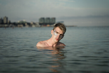 portrait of a non-binary young man with sparkles on his face, standing in the sea, in the background you can see the city