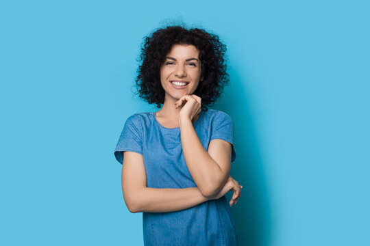 Caucasian Woman With Curly Hair Is Touching Her Chin And Smile On A Blue Wall Wearing A Summer Dress