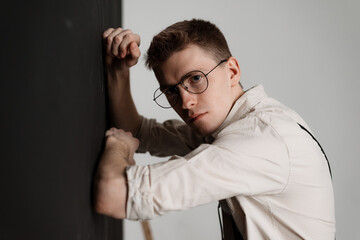 a young guy standing by the wall in a beige shirt and round glasses looking seriously at the camera