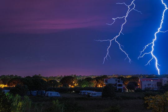 Two large and powerful lighting strikes crossing night blue sky and hitting urban area with houses and campervans in Zaton, Croatia