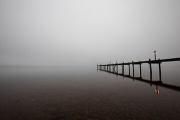 Fototapeta na wymiar Mole (pier) on the „Chiemsee“ lake in Bavaria, Germany. Wooden bridge in winter time with frozen and icy lake and dim lanterns for fishing. Dark and Foggy lake.