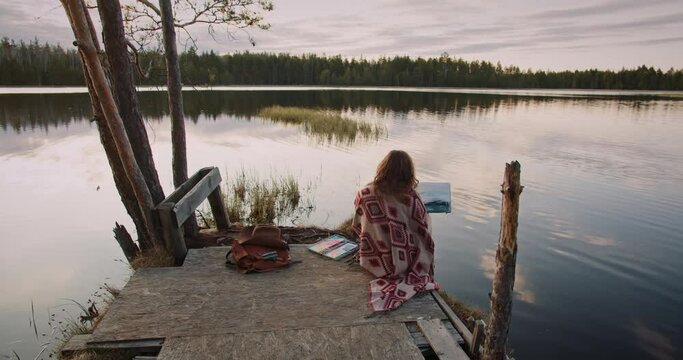A young artist girl sitting and drawing in front of forest lake with her sketchbook in watercolor technique. Outdoor wild nature painting concept