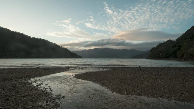 Time-lapse at sunset of Nikau Cove, Kenepuru Sound, Marlborough Sounds, New Zealand. A small stream running over the beach in the foreground with the tide advancing behind.