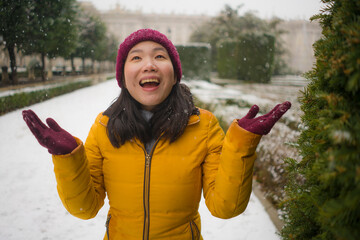 young happy and attractive Asian Korean woman in Winter jacket and beanie enjoying snowfall at city park playing cheerful surrounded by snow during Christmas holidays