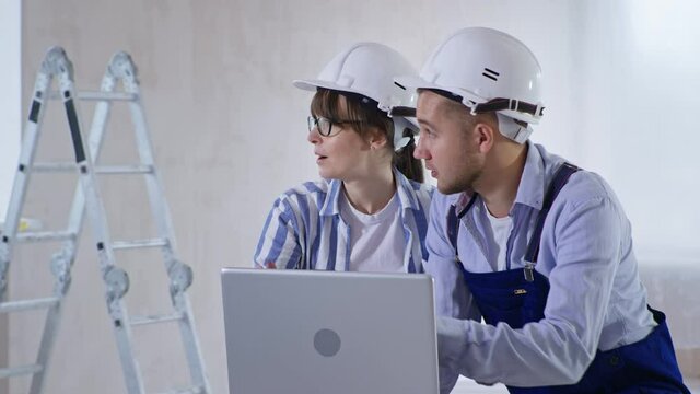 home improvement, young woman and man in helmets are discussing room renovation project on laptop and choosing wall color with color palette in their hands