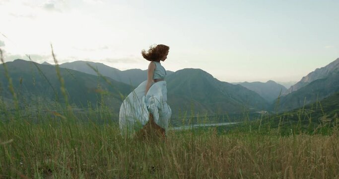Girl in light dress with red hair is walking on top of a spring mountain with scenic view. Woman in search for inspiration - freedom concept 4k footage