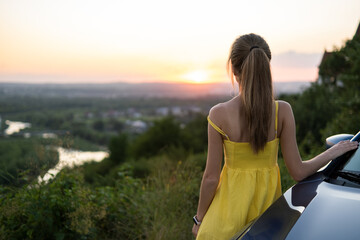 Carefree woman driver in yellow summer dress enjoying warm evening near her car.