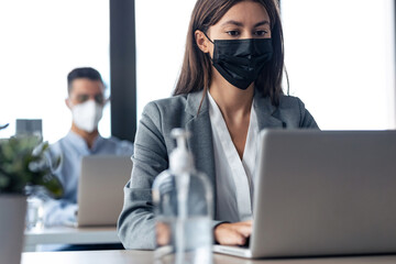 Two attractive business partners wearing a hygienic face mask while working with laptops in the coworking space. Social distancing concept.
