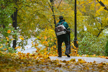 Seasonal work by city utilities in the park. A worker with a motorized backpack blower blows fallen...
