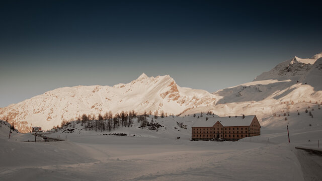 House of Simplon hospiz on the top of the simplon pass covered in winter snow. House surrounded with large amount of snow with visible mountains in the background bathing in sun.