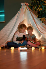 Two little kids, boy and girl telling stories to each other while sitting on a blanket in a teepee made with bedsheets at home © Svitlana