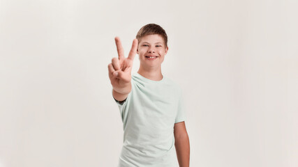 Happy disabled boy with Down syndrome smiling at camera, showing peace sign with one hand while...