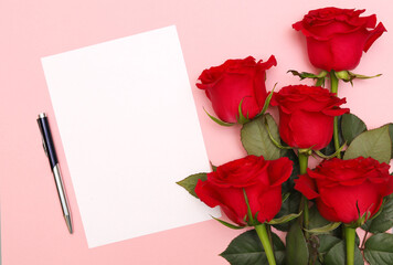 A bouquet of red roses and a blank white sheet of note paper with a pen on a pink background. Copy space. Concept: Gift for Valentine's Day, March 8, Mother's Day or birthday.The view from the top