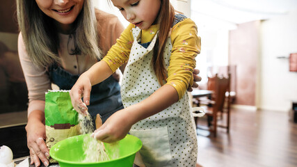Little girl pouring flour into large bowl beside grandmother