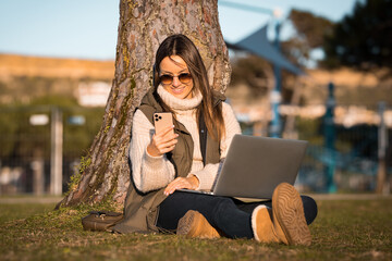 Photo of happy caucasian woman sitting on the grass outdoors in the park using laptop and mobile phone. Online and freelance work concept