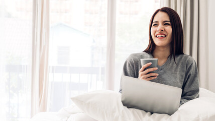 Portrait of smiling happy beautiful asian woman relaxing using technology of laptop computer while sitting on bed.Young creative girl working and typing on keyboard at home.work at home concept