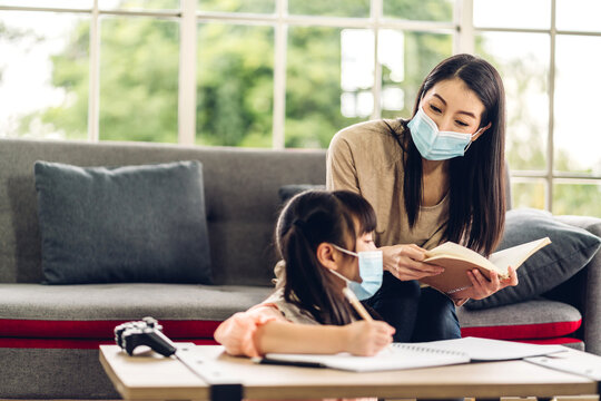 Portrait love asian family mother and little asian girl learning and writing in book with pencil making homework in quarantine for coronavirus wearing protective mask with social distancing at home