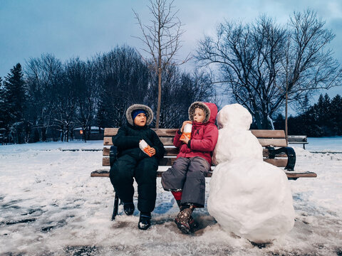 Two Cute Funny Children Friends Sitting On Bench In Park Outdoors On Winter Day Evening And Eating Cookies Snack. Kids Having Fun Outside And Relaxing After Making Snowman.