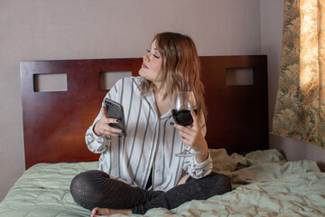 beautiful relaxed woman sitting on bed using cell phone with glass of red wine