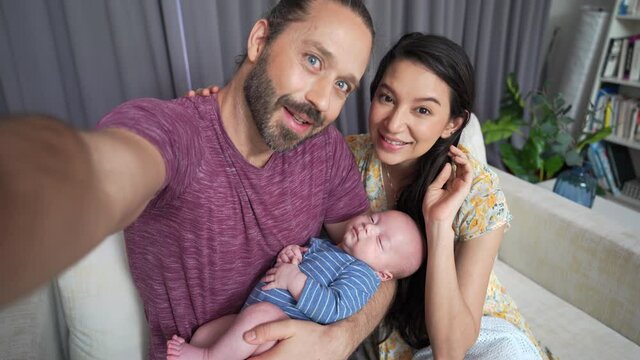 Caucasian man using smartphone taking selfie or home video of his family wife and adorable newborn baby son in living room. Father and mother relax and enjoy with infant child boy together at home.