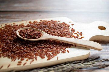 Red rice in a wooden spoon placed on a pile of red rice on a wooden tray and ears of rice on wooden table.