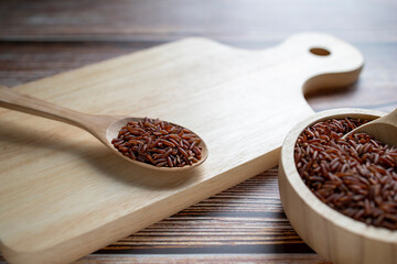 Red rice in wooden spoon on wooden cutting board and wooden bowl on table.