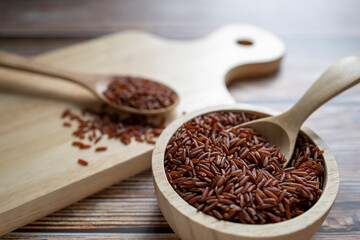 Red rice in wooden bowl with wooden spoon and red rice in wooden spoon on wooden cutting board on table.