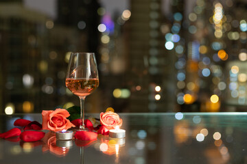 glass red wine, roses, petals and candles on table with reflection, twinkling blurred city night lights in background, romantic concept for lovers and valentine