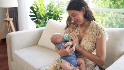 Happy family stay home. Beautiful Caucasian woman sitting on sofa in bedroom holding and feeding her adorable newborn baby son. Sleepy cute infant child boy drinking milk from bottle on mother arm.