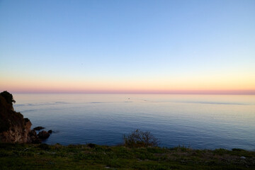 View from a high cliff on water of sea and pink and yellow sunset on horizon in background