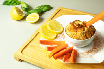 A bowl of hummus with paprika. Some carrot sticks are placed next to the bowl and one is dipping into the spread. Lemon slices and basil. Healthy vegeterian and vegan meal, appetizer or snack.