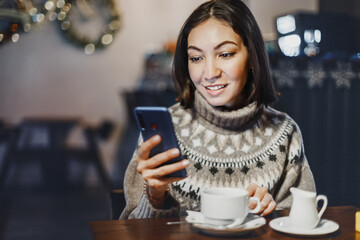 A young woman copywriter or blogger works with social networks on her smartphone while sitting in a coffee shop and sipping hot coffee