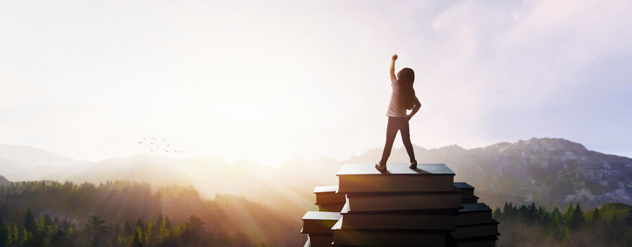 concept - little girl with raised fist on giant books at the top of the mountain in beautiful sunset