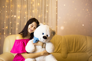 Happy girl in a pink dress hugging a big white soft bear home on the couch. Gift from a loved one, valentine's day, birthday, holiday. Copy space