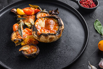 Served portobello  mushrooms,baked and stuffed with cheddar cheese, cherry tomatoes and sage on black plate over  black background side view.