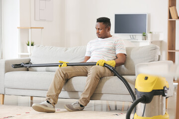 African-American man with vacuum cleaner sitting on sofa