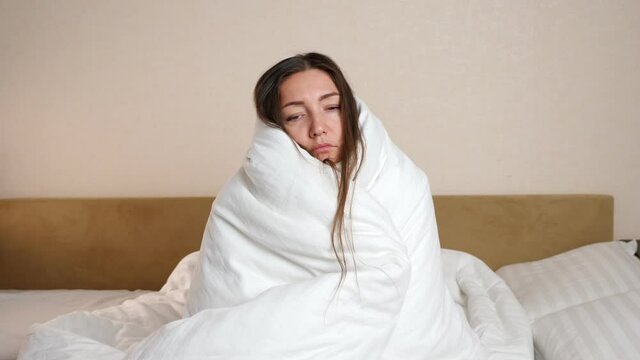 Sick woman wrapped with soft white duvet sits on large bed in bedroom