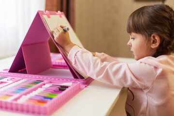 Child drawing with color wax crayons, side view of concentrated little girl with pigtails paints, using drawing set for painting and art, female kid wearing casual shirt sitting at table.