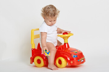 Little boy sit on red toy car and holding tiny vehicle in hands, looking on floor, barefoot child wearing bodysuit playing isolated over white background.