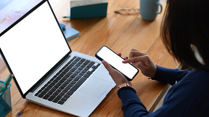 Cropped shot of young female sitting in front of her laptop computer and using smart phone.