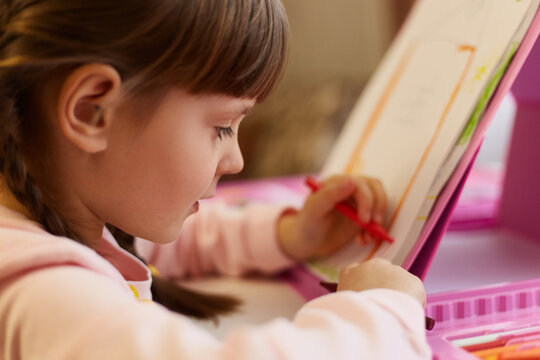 Little girl painting on paper, using red wax crayon, looking concentrated on paper, tries to draw beautifully, sitting at table, female kid with dark hair.