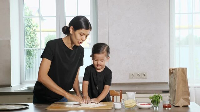 Mother and little daughter wearing same black shirts stir and stretch elastic dough for pizza above wooden cutting board poured with flour