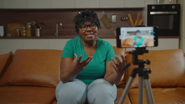 Positive experienced mature black woman web influencer in eyeglasses sitting on cozy couch at home, recording content for online vlog on smart phone, talking about happy retirement with followers.