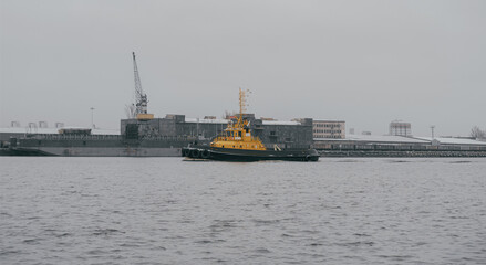 Fototapeta premium Tugboat sailing on the river. Gloomy, bad weather, gray sky, port in the background.