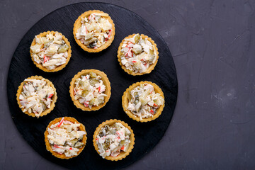 Tartlets with salad on a black wooden round board laid out in a circle on a gray concrete background. Copy space